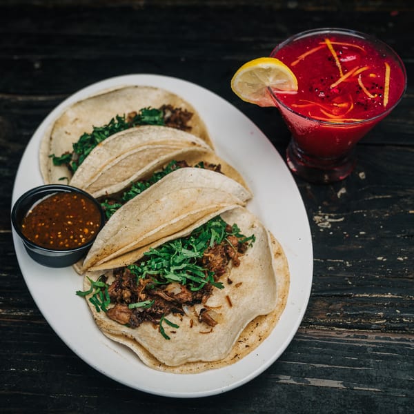 Three Salty Joe's BBQ tacos with salsa and a red drink with lemon on a dark wooden table.