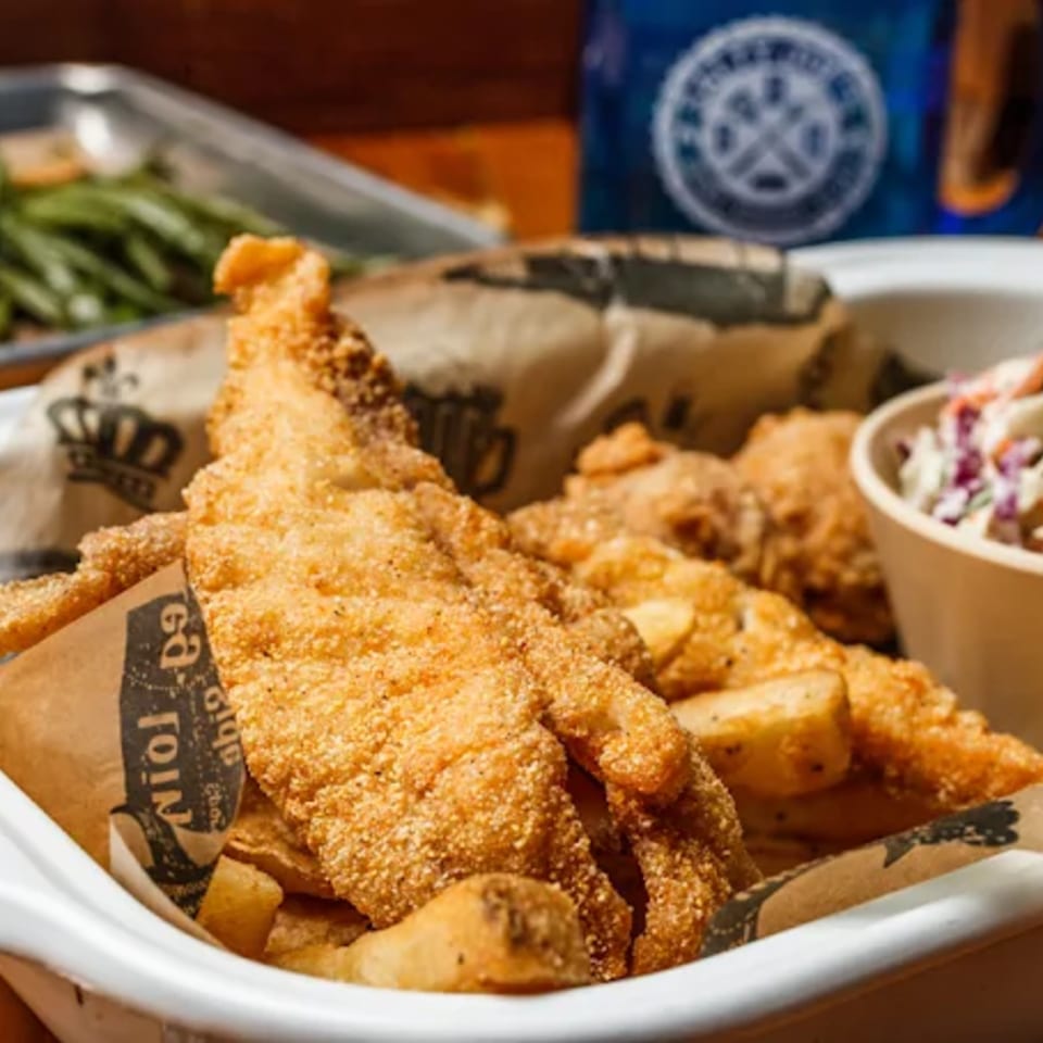 Crispy fried fish and fries in a white container with coleslaw and green beans in the background.