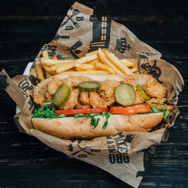 Salty Joe's BBQ fried shrimp po'boy with pickles, lettuce, tomatoes, and fries on branded paper.