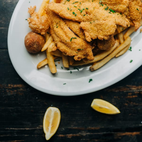 Crispy fried fish fillets, hushpuppies, and french fries on a white plate with lemon wedges.