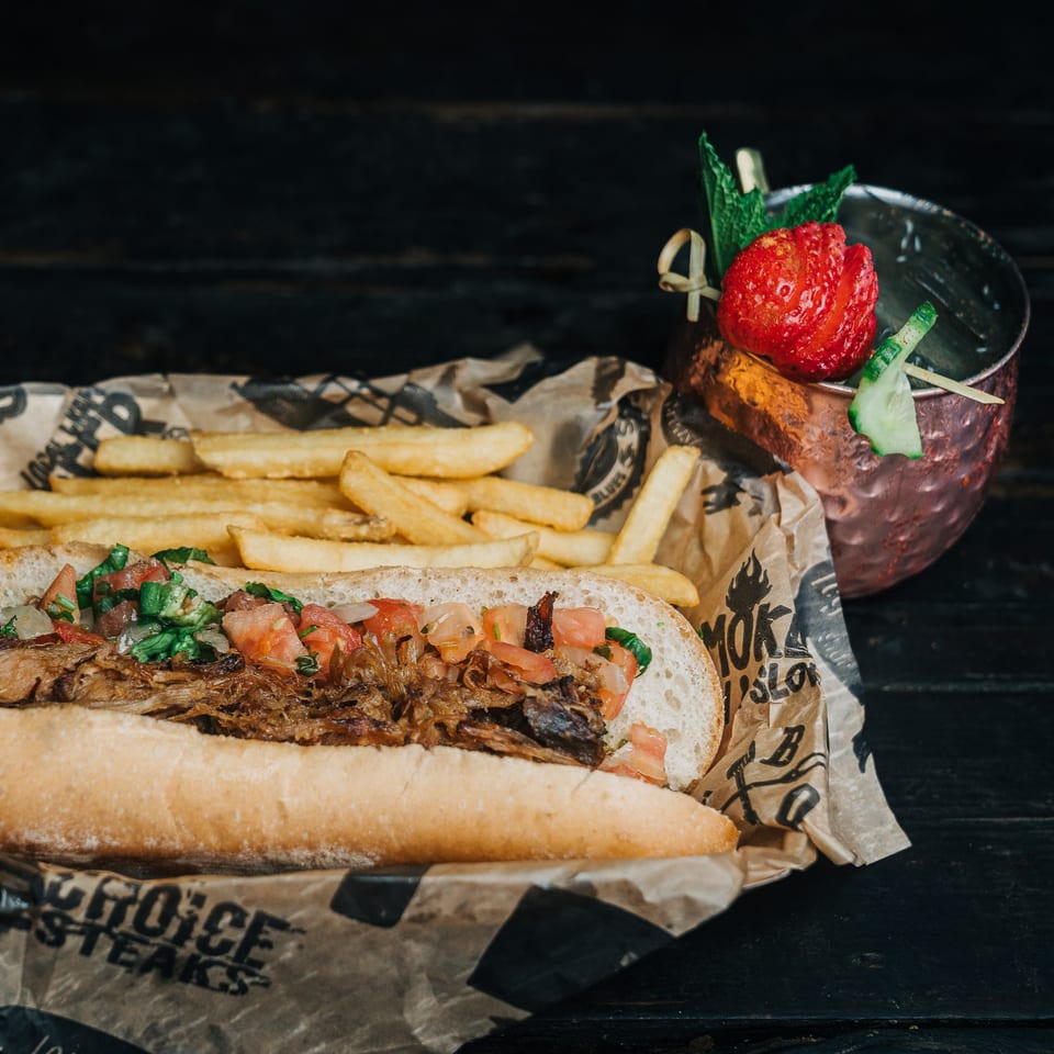Salty Joe's BBQ pulled pork sandwich with fries and a garnished cocktail on a dark wooden table.