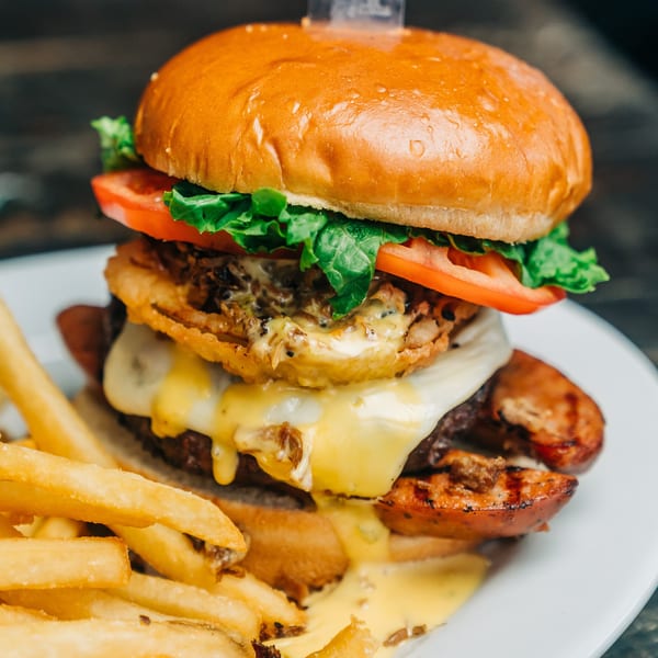 A towering Salty Joe's BBQ burger with melted cheese, onion rings, and fries on a white plate.