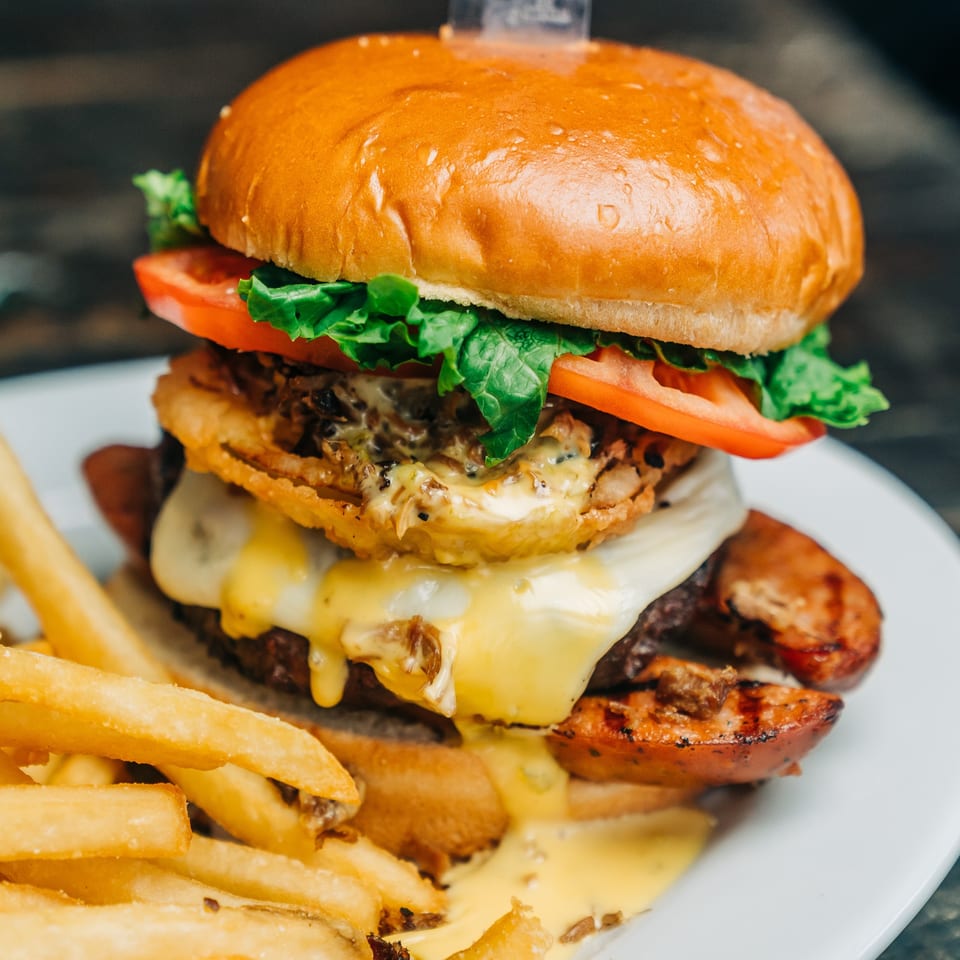 A towering Salty Joe's BBQ burger with melted cheese, onion rings, and fries on a white plate.