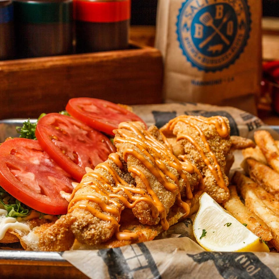 Salty Joe's BBQ fried fish sandwich with tomatoes, lettuce, fries, and a lemon wedge.